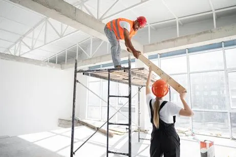 Um homem e uma mulher na construção; Homem estã em cima de um andaime e a mulher embaixo ambos estão segurando uma tábua